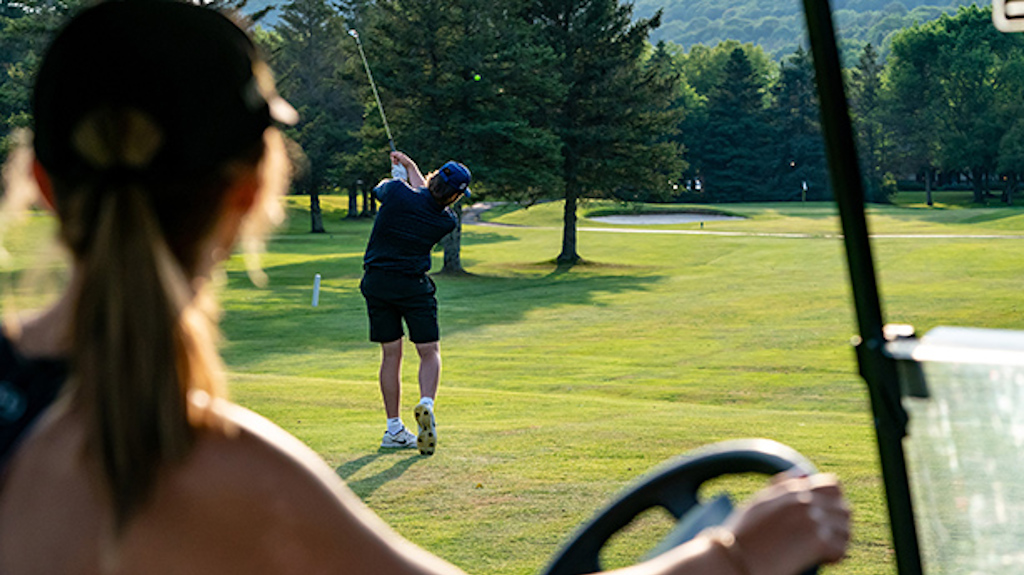A young woman sits in a golf cart while watching a man tee of on the Double Black Diamond Golf course.